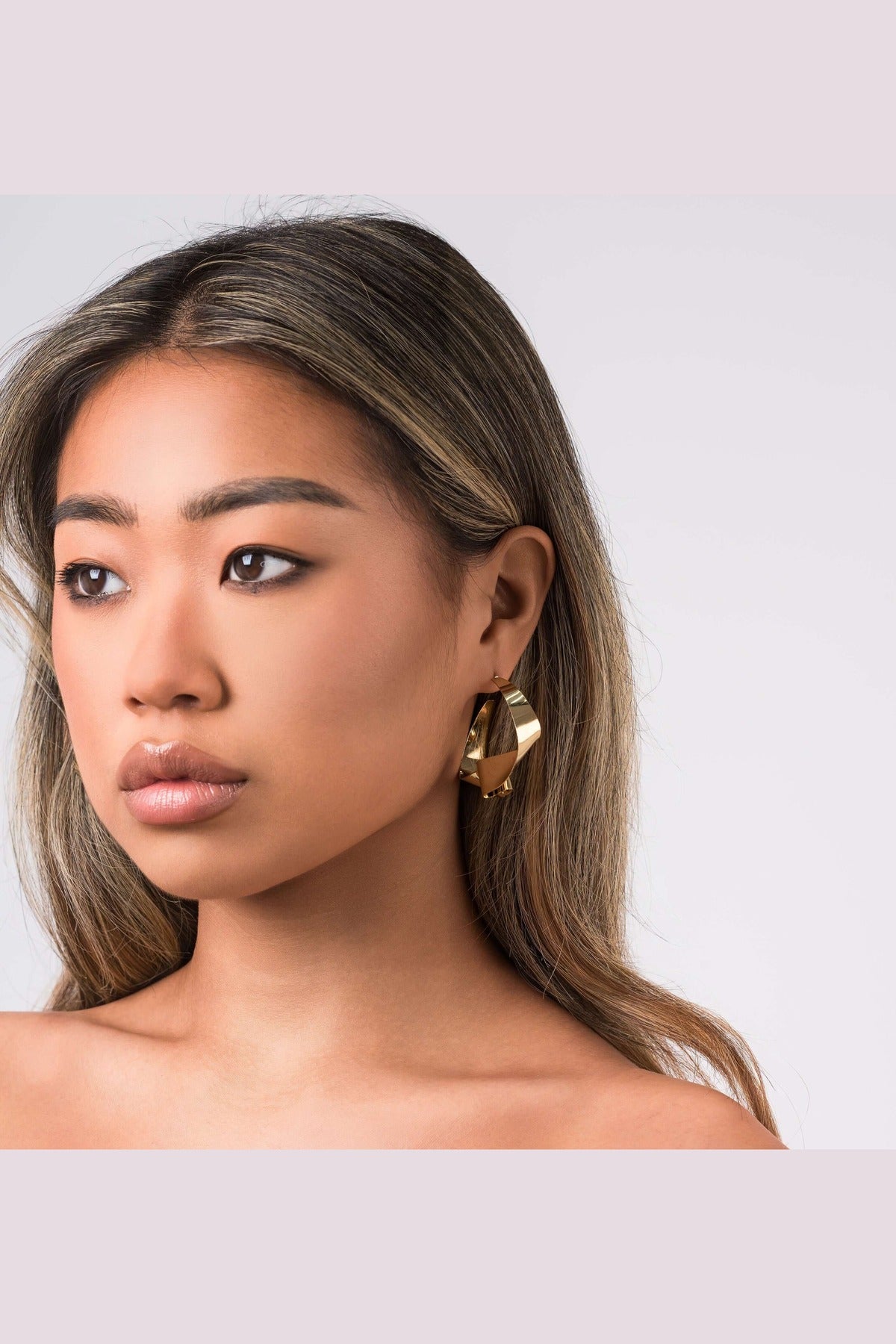 A model showcasing ARIIRA Alma Folded Ribbon Hoop Earrings, featuring a sculptural gold design, elegant and lightweight.